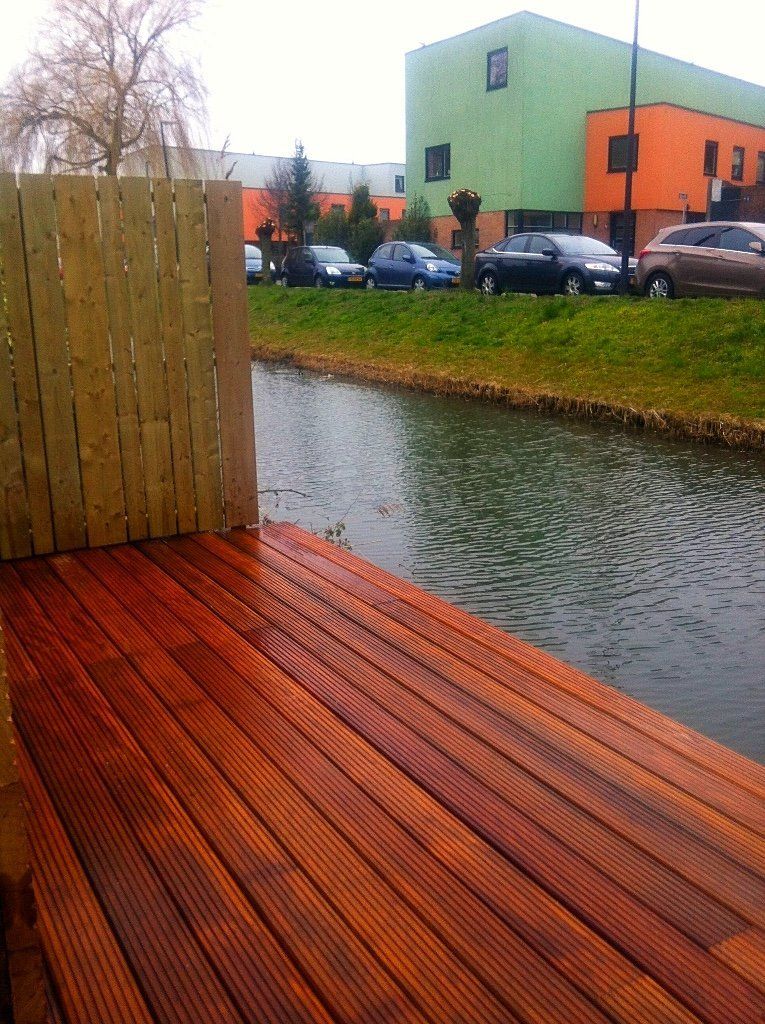 Houten terras naast een kanaal. Kleurrijke gebouwen op de achtergrond, auto's geparkeerd aan de zijkant.