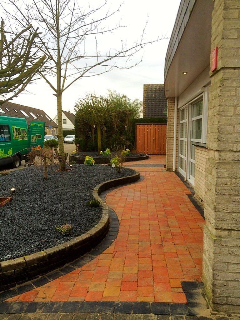Een bakstenen pad kronkelt langs een gebouw en een donkerstenen tuinperk met een kale boom.