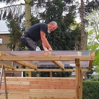 Man op het dak van een schuur, bezig met het installeren van transparante dakpanelen. Buiten, bewolkte dag.