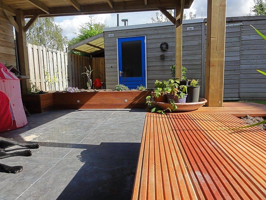 Buitenruimte met houten vlonder, grijze patio, schuur met blauwe deur en planten.