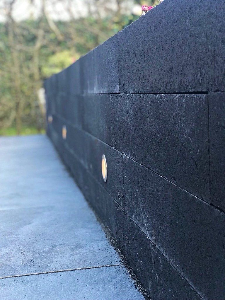 Zwarte, rechthoekige keermuur van blokken met ingebouwde verlichting, tegen een wazige buitenachtergrond.