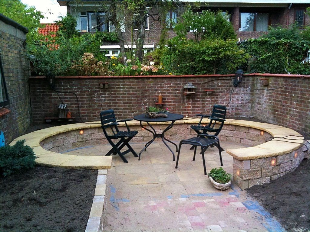 Terras met een ronde stenen bank, zwarte stoelen en een tafel in een tuin met bakstenen muren.