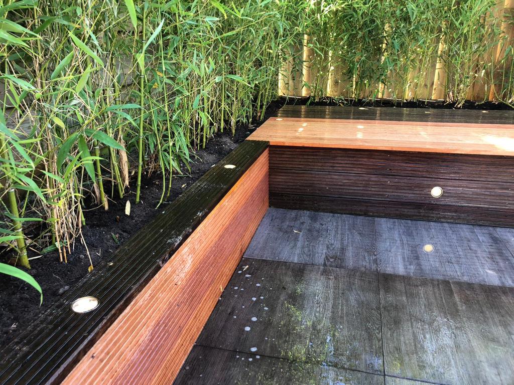 Houten bloembed met ingebouwde verlichting, naast bamboeplanten en een betegeld terras.
