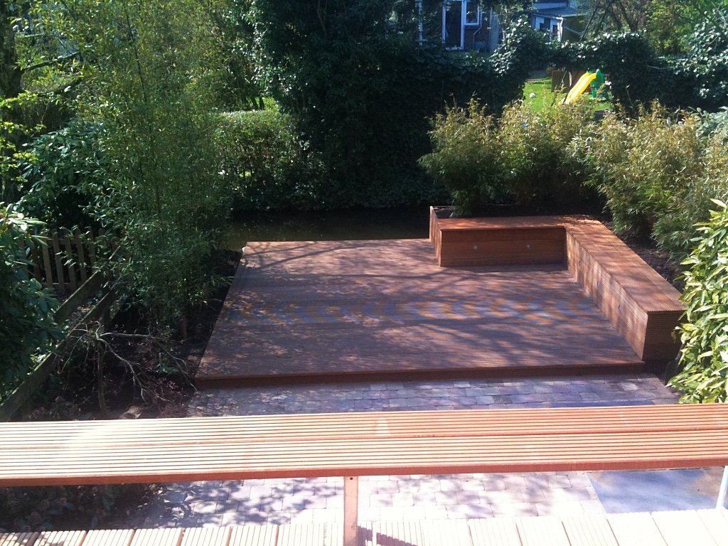 Houten terras met ingebouwde bank in een tuin, omgeven door groen.