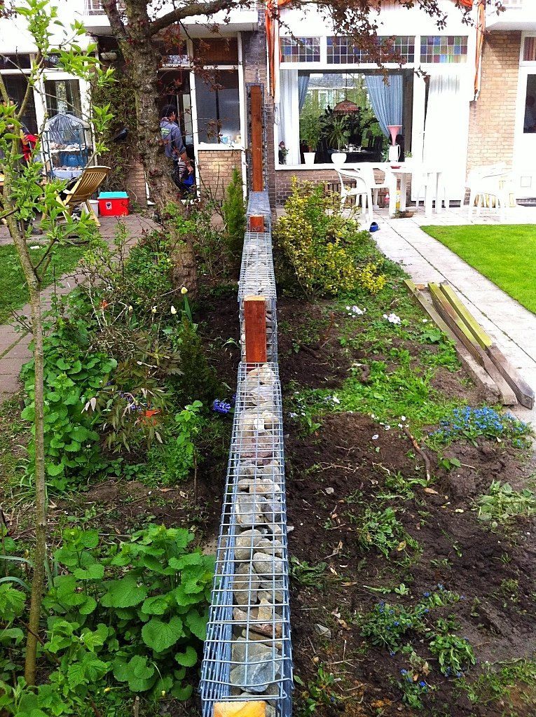 Een met stenen gevulde draadkooi als keermuur in een achtertuin, met planten en een glimp van het huis.