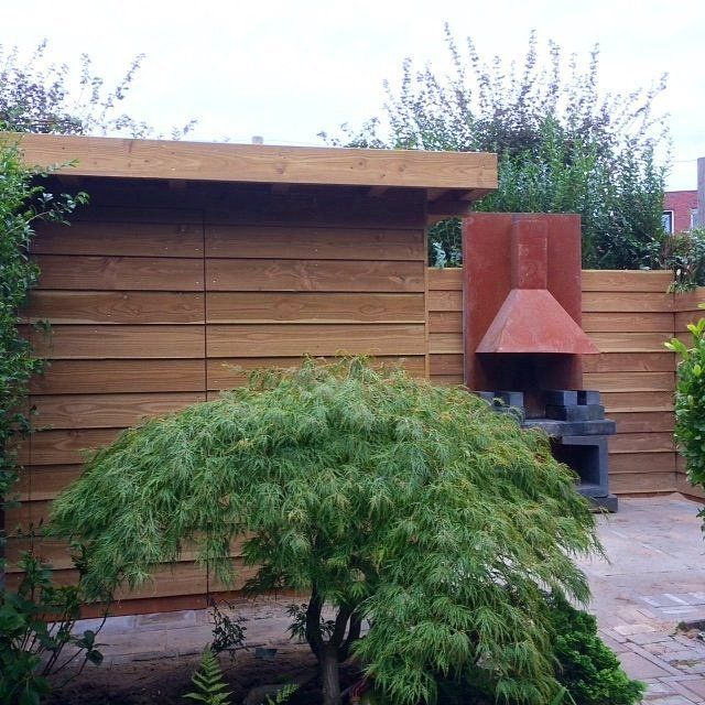 Houten buitenconstructie met grill, afgedekt door een metalen kap. Op de voorgrond staat een treurboom.