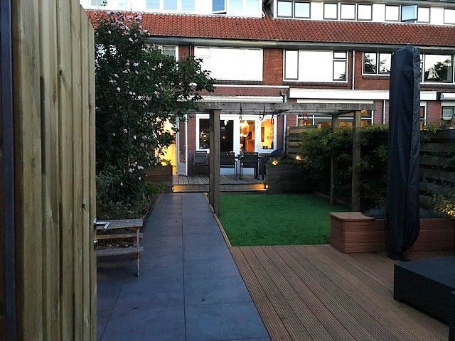 Achtertuin met houten terras, looppad en pergola die naar een huis met ramen leidt. Groen gras.