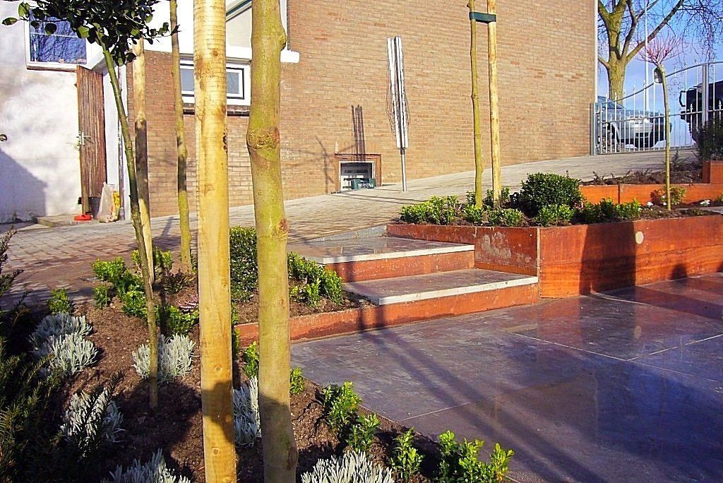 Buitenruimte met verhoogde houten plantenbakken, trappen en jonge bomen. Op de achtergrond een bakstenen gebouw.