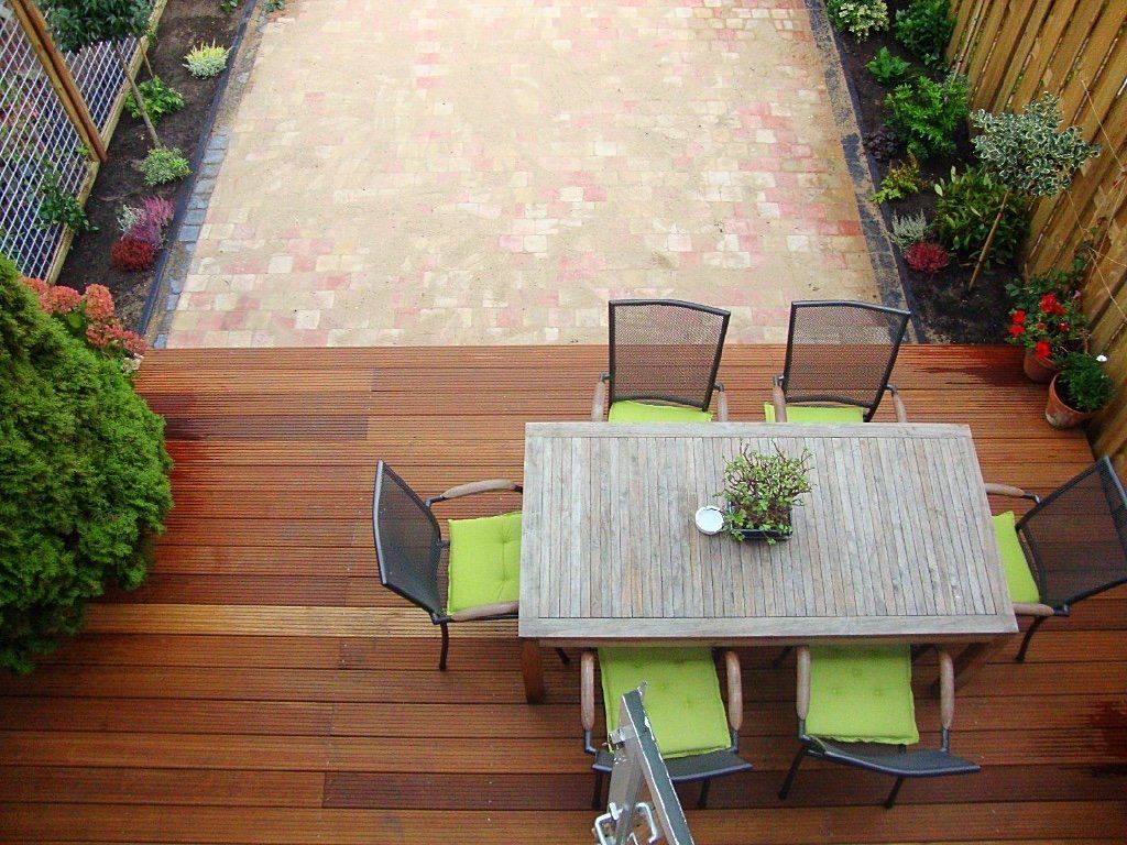 Houten terras met tafel en stoelen, een stenen patio en omringende beplanting.