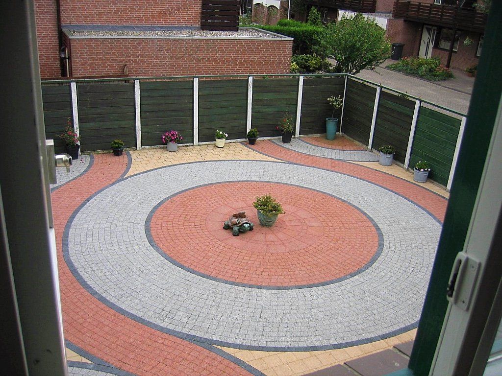 Rond terras van bakstenen in een achtertuin, omgeven door hekken en paden. Bakstenen in rode, grijze en gele kleuren.