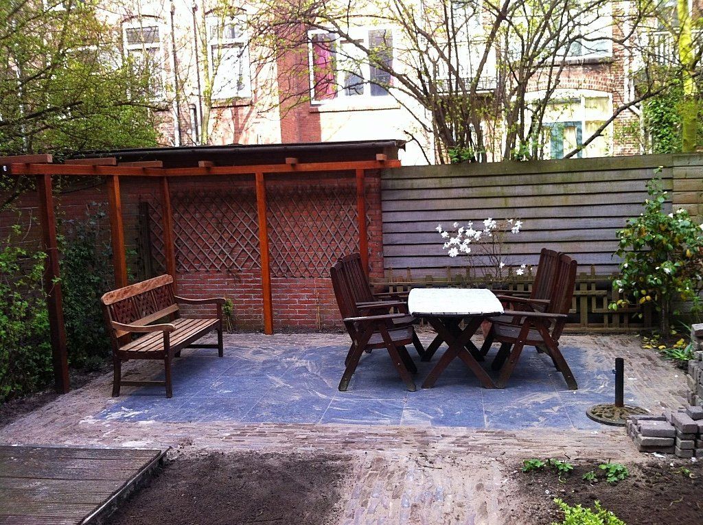 Terras in de achtertuin met houten tuinmeubelen en een overkapping; op de achtergrond een bakstenen muur en een schutting.
