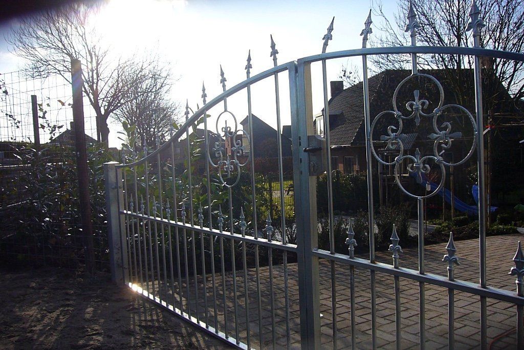 Metalen poort met sierlijke leliemotieven, die op een zonnige dag toegang biedt tot een tuin en huizen.