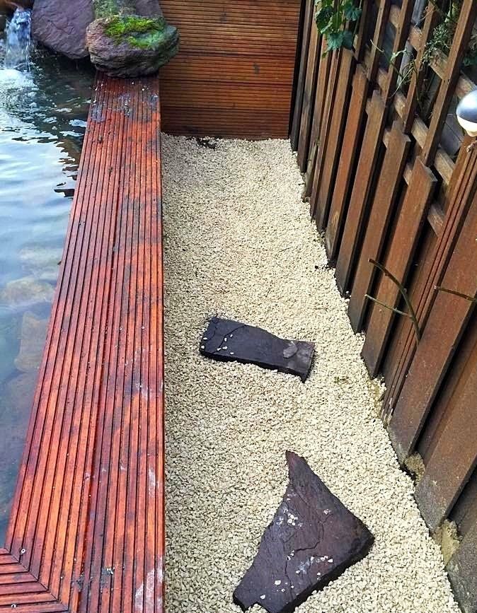 Houten terras en schutting langs een grindpad met stapstenen naast een waterpartij.