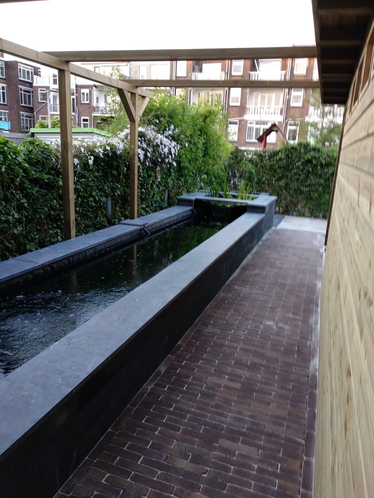 Een langwerpige, rechthoekige vijver met zwarte rand en een bakstenen pad, onder een houten pergola, vlakbij een houten gebouw.