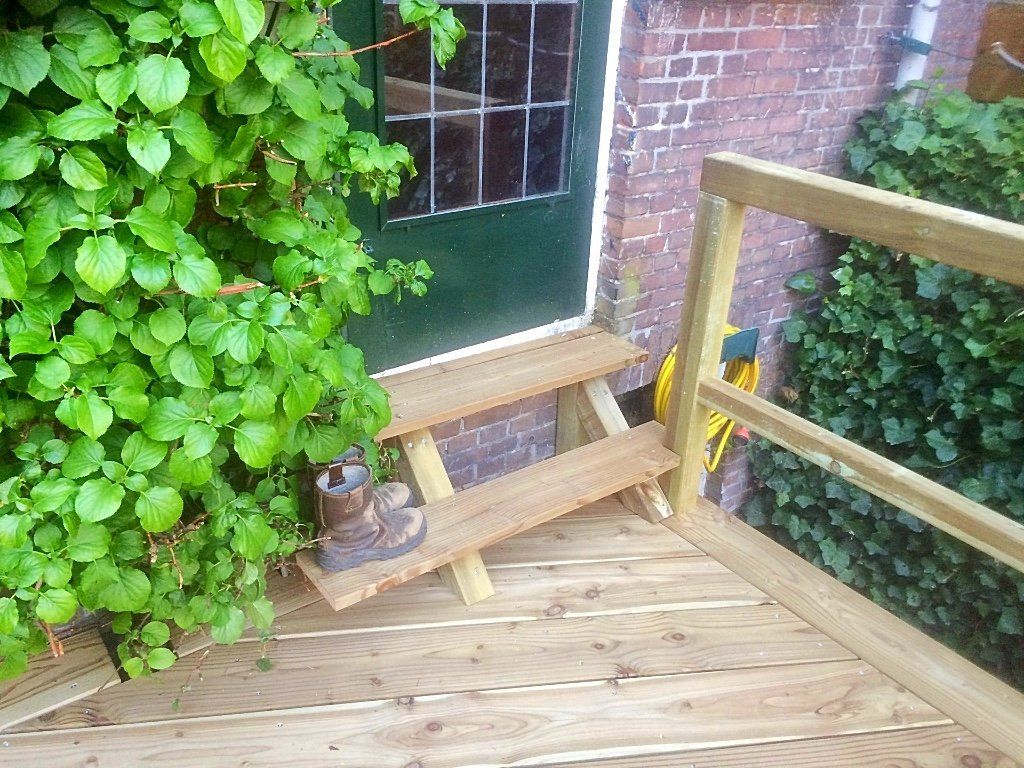 Houten treden die leiden naar een groene deur, met een houten leuning en omringende begroeiing.