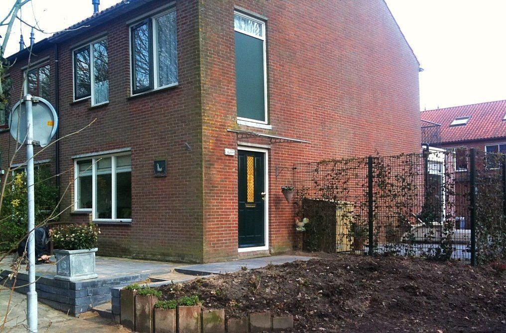 Een bakstenen huis met twee verdiepingen, een groene deur, een groen raam en een kleine tuin met een hek.
