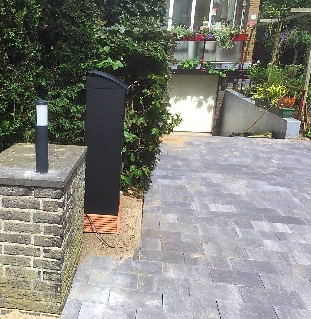 Zwarte buitenluidspreker naast een bakstenen pilaar en een betegeld terras, met een groene heg en een garagedeur op de achtergrond.