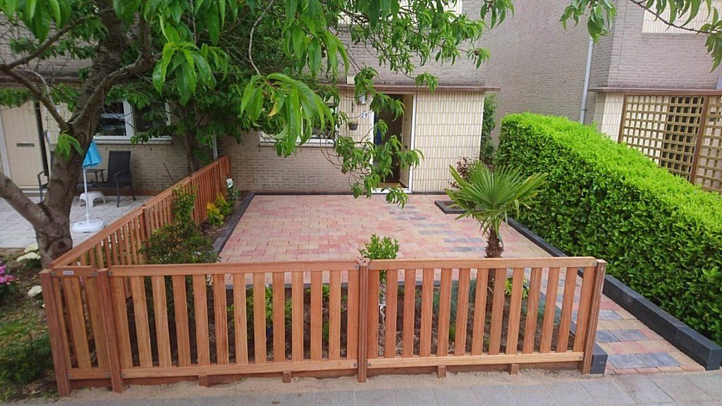 Een houten schutting omsluit een kleine tuin met een betegeld terras, bomen en hagen voor een gebouw.