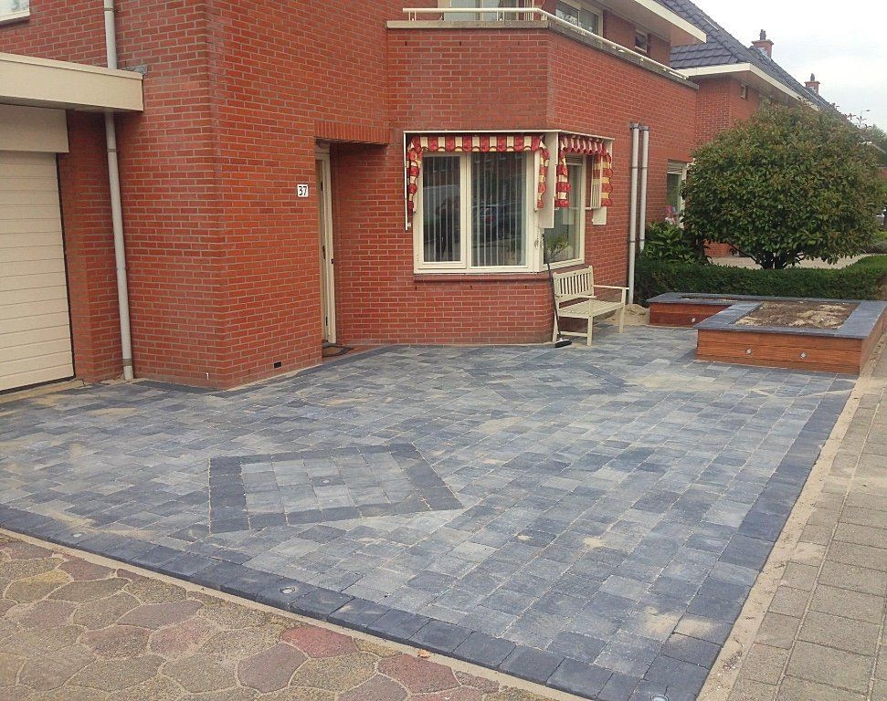 De buitenkant van een bakstenen huis met een blauwgrijze oprit, een rode bakstenen muur, een klein raam met een luifel en een kleine boom.