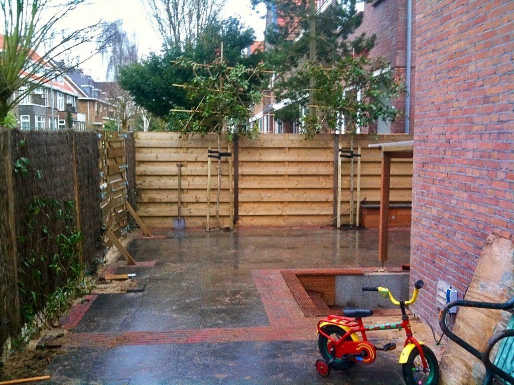 Achtertuin met houten schutting, bakstenen muur, natte bestrating en een driewieler.