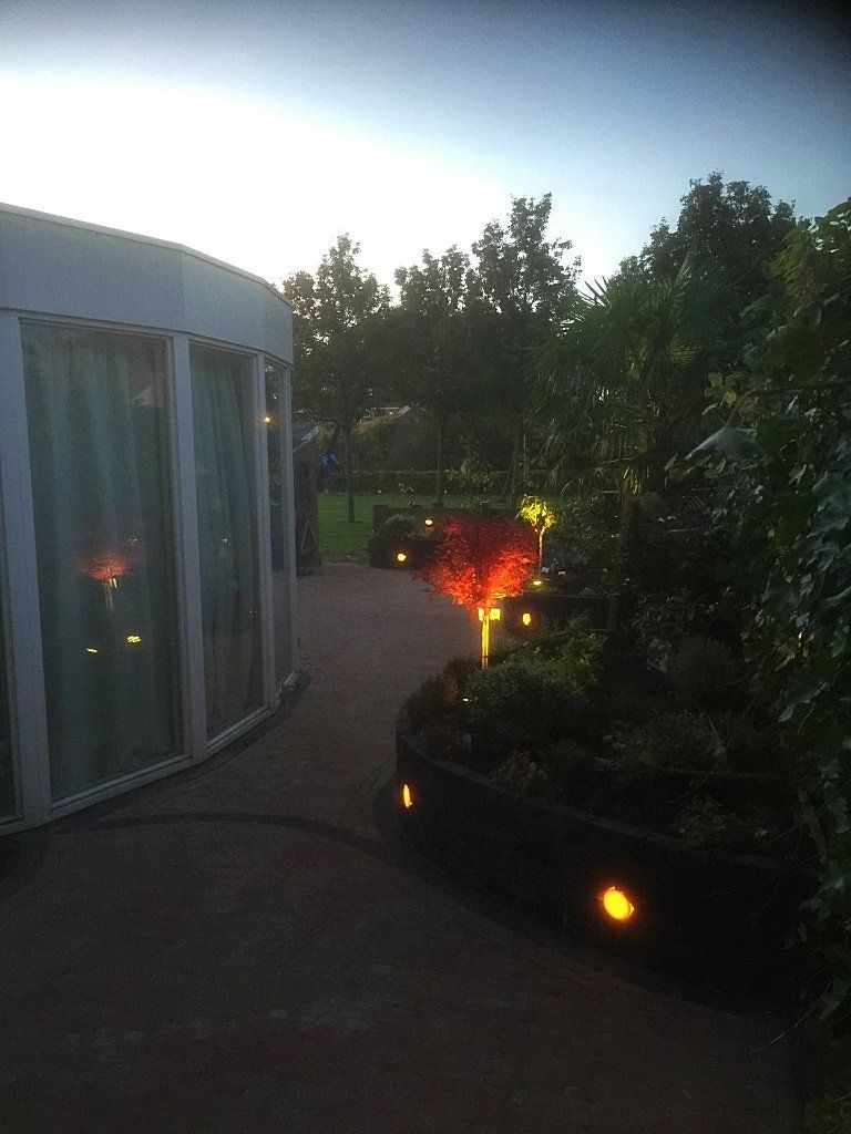 Een gebogen wandelpad met tuinverlichting, langs een gebouw met glazen wanden, in de schemering.