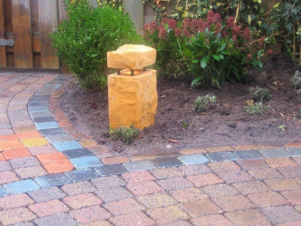 Stenen sculptuur naast een bakstenen pad en een bloembed met groen.