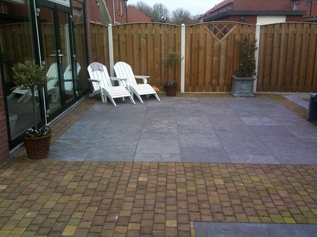 Terras met twee witte stoelen, planten en een houten schutting. Grijze en baksteenkleurige bestrating.