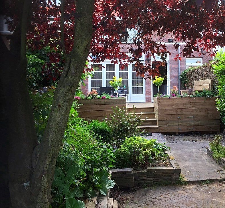 Tuin met houten plantenbakken en een trap die leidt naar een bakstenen huis met witte deuren, onder een boom met rode bladeren.