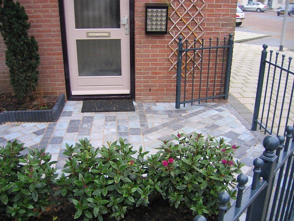 Ingang van een bakstenen gebouw met roze deur, zwarte poort, sierbestrating en struiken.