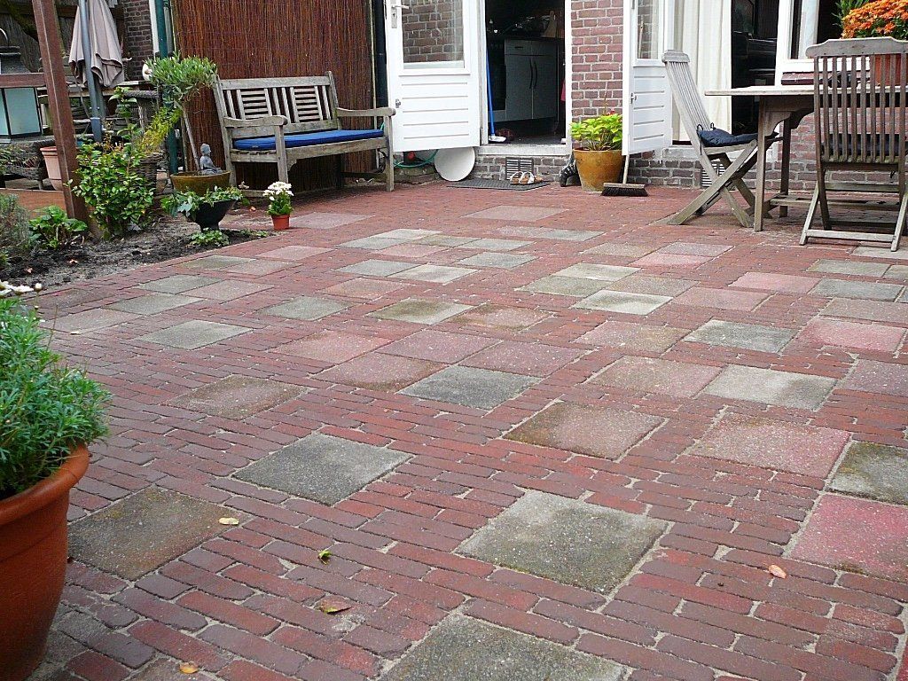 Een stenen terras met afwisselend vierkante en rechthoekige bestrating in een achtertuin; deur en bankje zichtbaar.