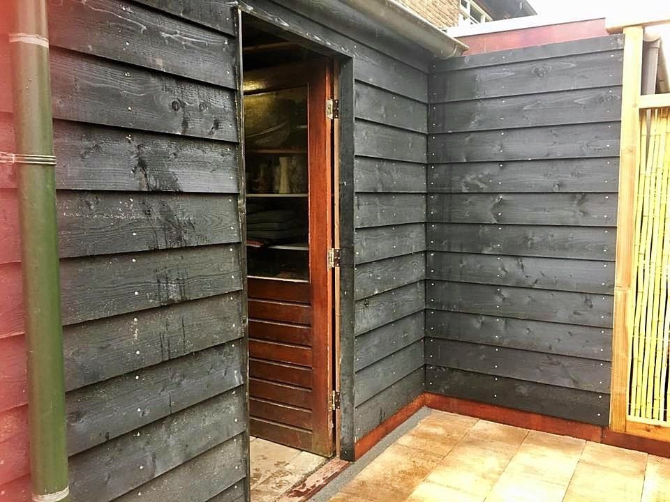 Zwarte houten schuur met een open deur naar een schemerig verlicht interieur. Aangrenzende muur met een geel tralieraam.
