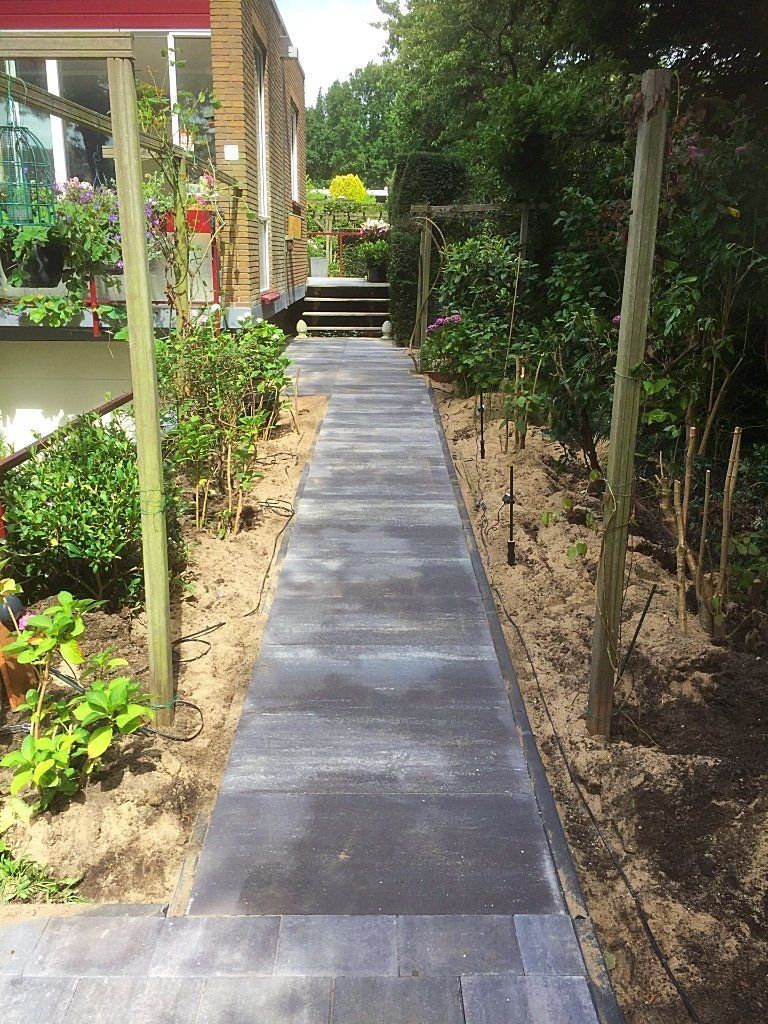 Een stenen pad loopt tussen bloembedden met planten en houten palen door naar een gebouw.