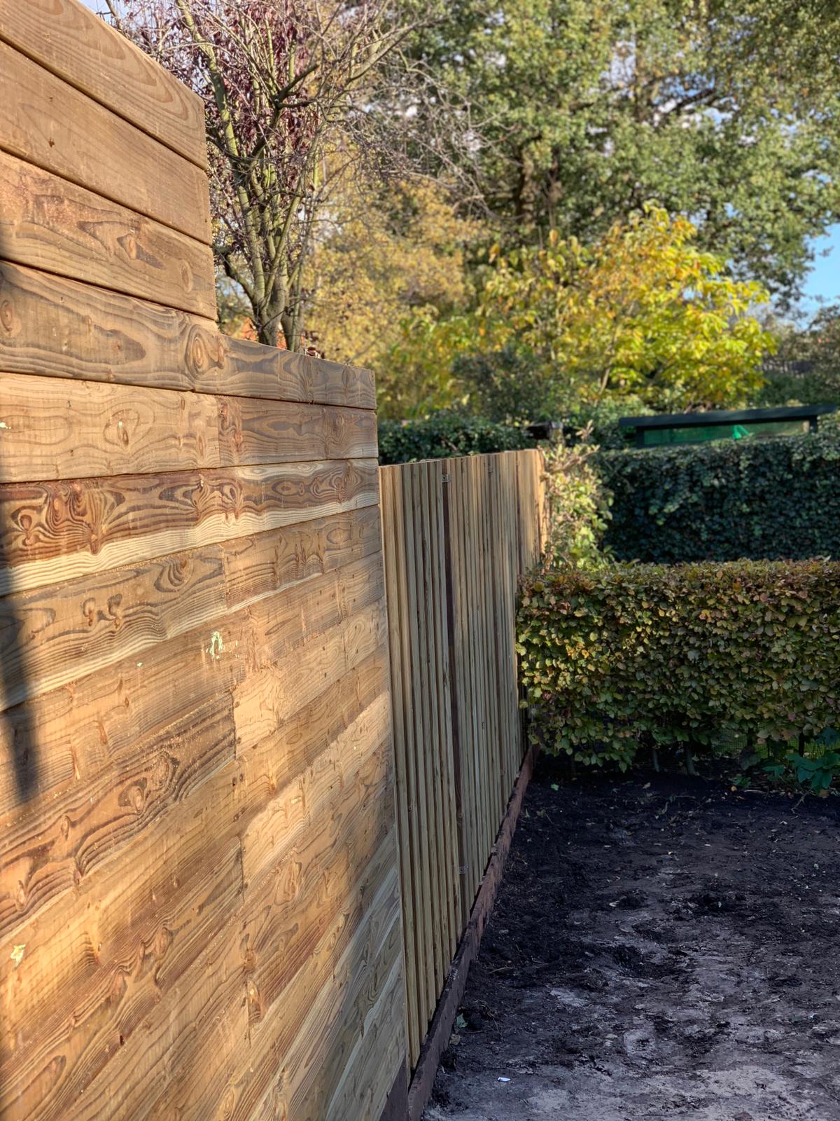 Hoek van een houten schutting in een tuin, zonlicht. Bruin hout, groene heg, bomen op de achtergrond.