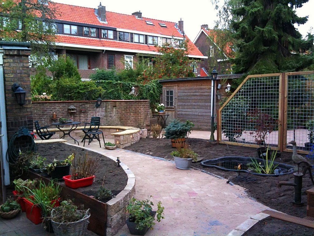 Een achtertuin met een kronkelend pad, een kleine vijver, een zitgedeelte en een houten pergola. Op de achtergrond zijn bakstenen muren en gebouwen met rode daken te zien.