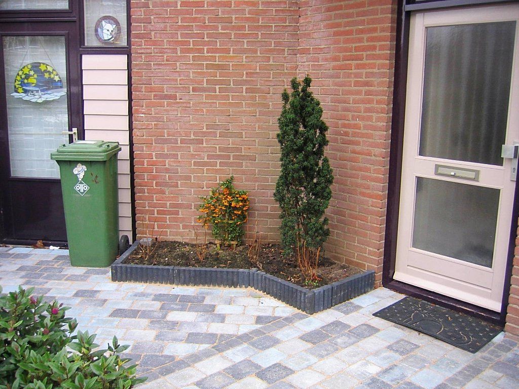 Een bakstenen muur met een kleine plantenbak met twee struiken ernaast, naast een voordeur met een deurmat. Links staat een groene vuilnisbak.
