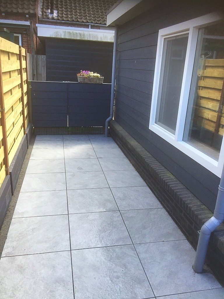 Smal terras, geplaveid met grijze tegels; donkerblauw gebouw met wit omlijst raam.