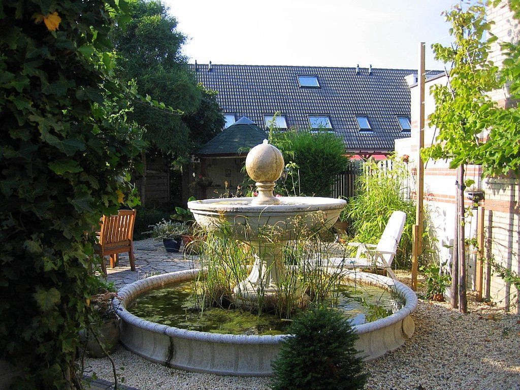 Fontein op een binnenplaats met groen eromheen, een tafeltje en stoelen, en gebouwen op de achtergrond.