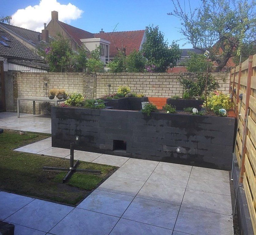 Achtertuin met een donkergrijze plantenbak gevuld met bloemen, een betonnen terras, gras en een houten schutting.