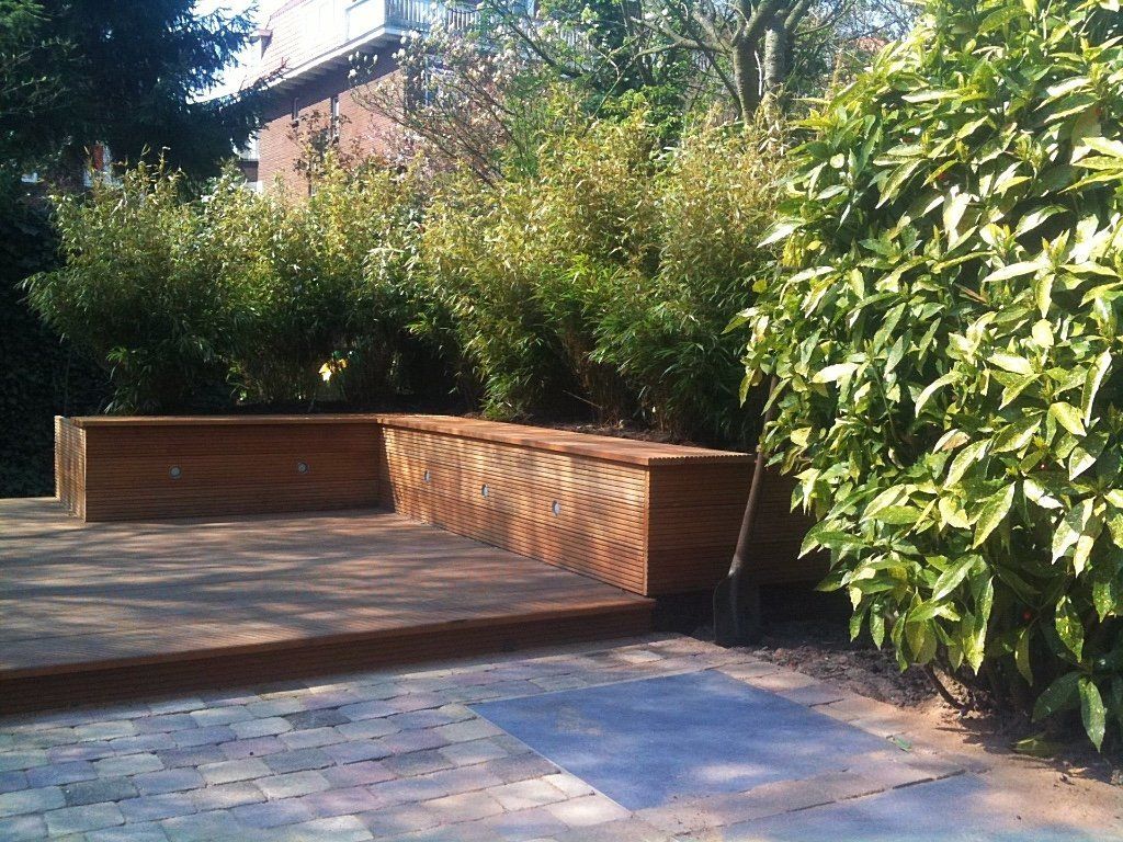 Houten platform met ingebouwde bank, omgeven door begroeiing en bakstenen bestrating.