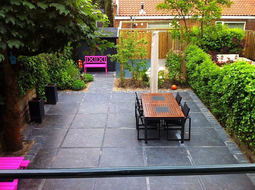 Terras met donkergrijze bestrating, een houten tafel met stoelen en weelderige groene hagen.