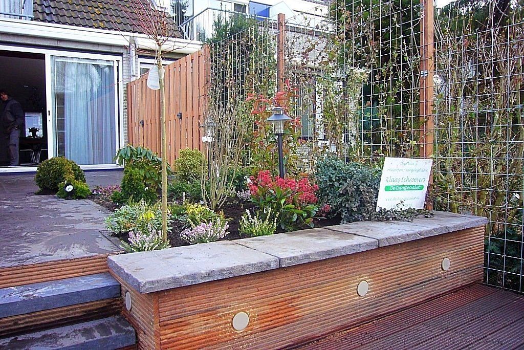 Bloembed met diverse planten, een houten schutting en klimplanten. Stenen bank op de voorgrond.