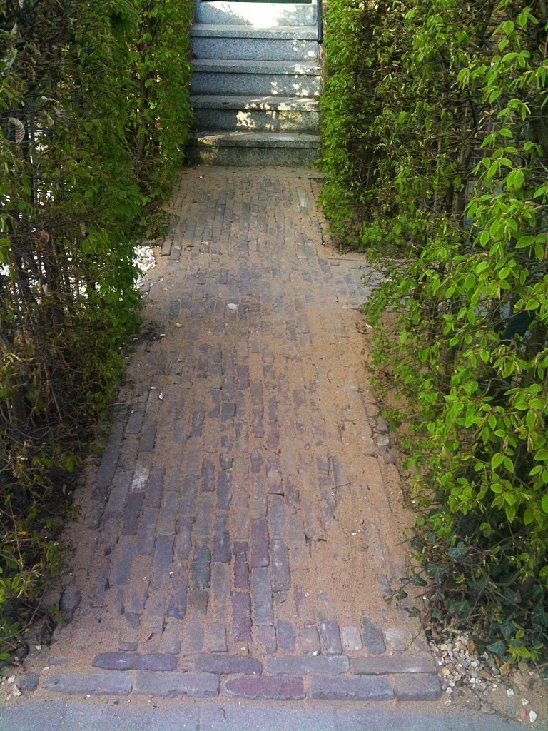 Een bakstenen pad, geflankeerd door groene hagen, leidt naar een trap en een poort.