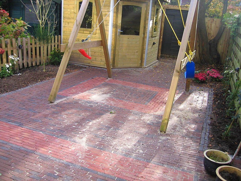 Speeltuin met bakstenen bestrating, schommel en houten speelhuisje in een achtertuin.