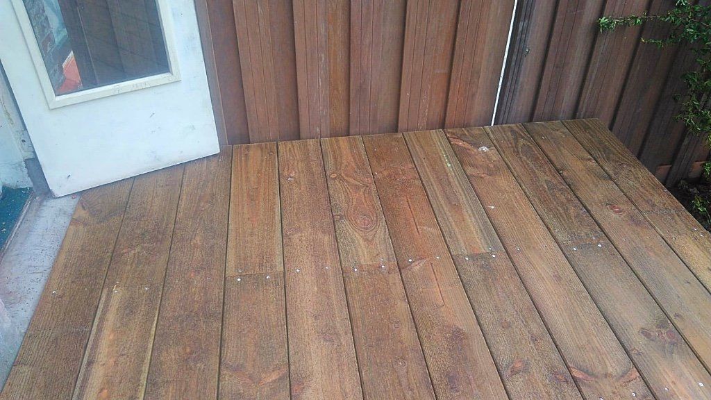 Een houten terras naast een verweerde houten schutting en een witte deur met een raam.