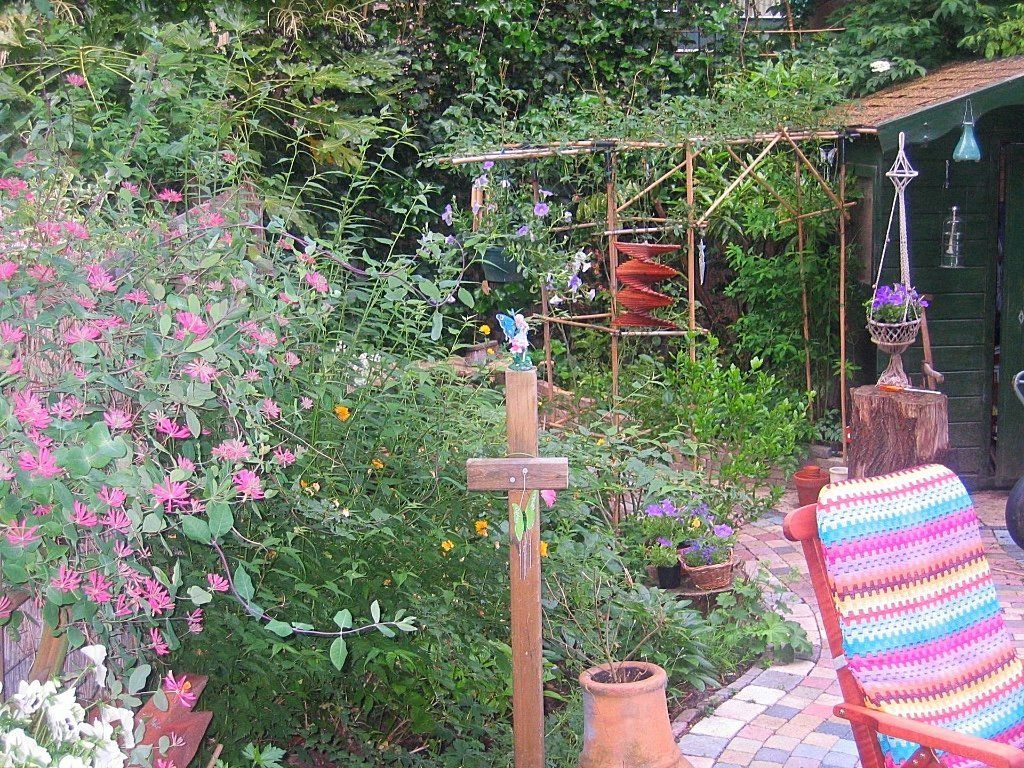 Een weelderig tuintafereel met roze bloemen, een houten vogelvoederhuisje en een kleurrijke gehaakte stoel op een stenen terras.