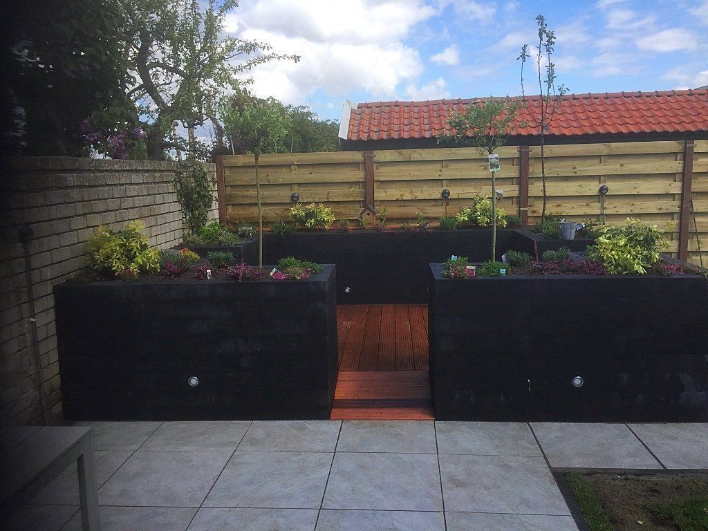 Zwarte, verhoogde bloembedden met planten en een looppad op een betegeld terras, met een houten schutting op de achtergrond.