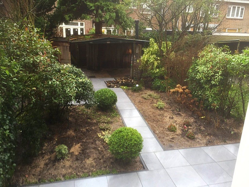 Tuin met betegeld pad, groene struiken en een houten schuur op de achtergrond.