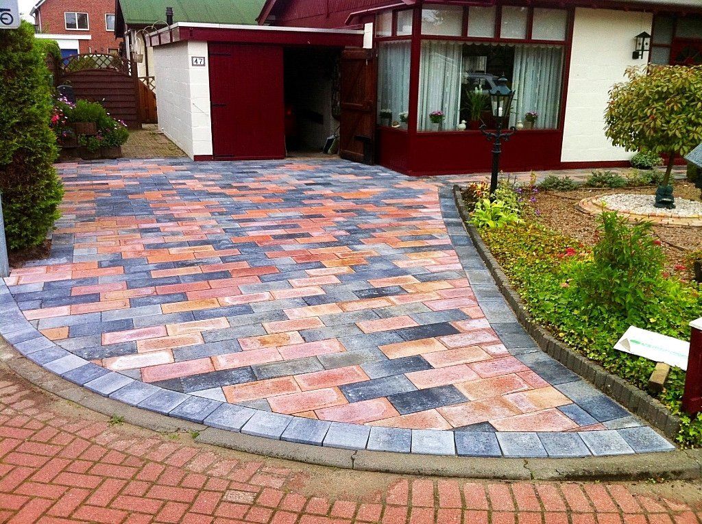 Oprit geplaveid met veelkleurige bakstenen, omzoomd door grijze bestrating en een kleine tuin, die leidt naar een huis met rode accenten.