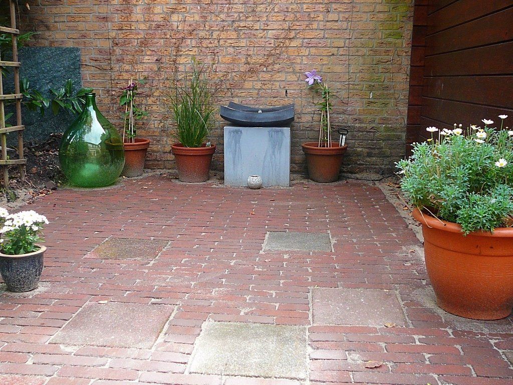 Een stenen terras met potplanten tegen een stenen muur. Een grote groene glazen fles en een stenen doos zijn zichtbaar.