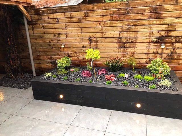Een zwarte, verhoogde plantenbak met planten, zwarte stenen en verlichting, tegen een houten schutting.